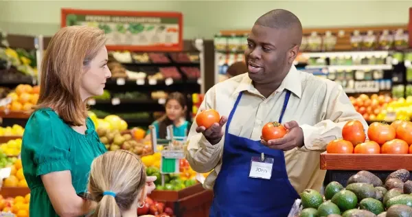 Grocery store worker handling tomatoes and talking to customer