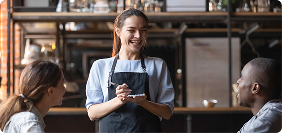 Smiling restaurant server in an apron taking an order from two customers at a table